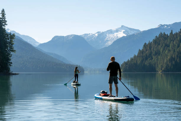 Couple paddle SUP boards across mountain lake, Cheakamus Lake, Whistler